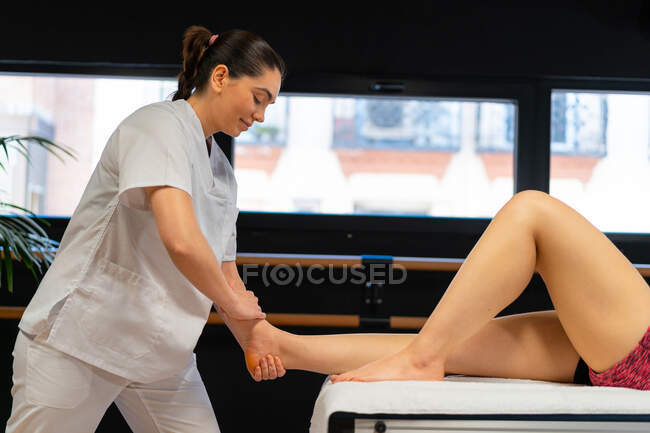 Seitenansicht der glücklichen Masseurin im weißen Gewand, die das Kalb einer Patientin während einer Physiotherapie-Sitzung in der Klinik massiert — Stockfoto