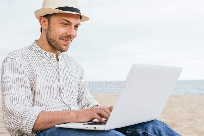 Vista lateral do jovem blogueiro masculino em uso elegante e chapéu sentado na praia de areia e digitando no laptop durante as férias de verão na costa — Fotografia de Stock