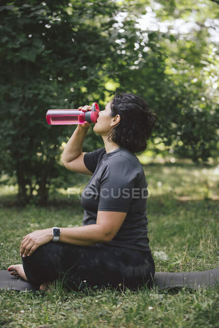 Вид збоку втомленої молодої леді з темним волоссям у спортивному одязі, що сидить на килимку на трав'янистому газоні з схрещеними ногами та питною водою після тренування йоги в парку — стокове фото