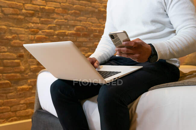 Low angle of crop ethnic male entrepreneur sitting on bed and browsing mobile phone and working on laptop — Stock Photo