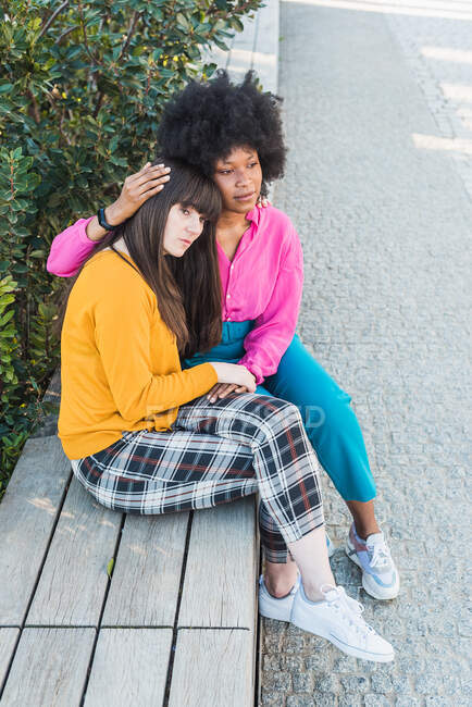 High angle of tranquil multiracial lesbian couple sitting on bench and embracing in city while looking away — Stock Photo