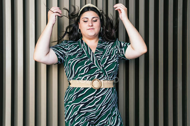 Stylish young plus size female wearing elegant dress with geometric diagonal ornament and belt standing with hands raised against striped wall — Stock Photo