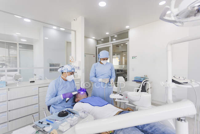 Женщина врач носить медицинскую форму и маску исцеляющие зубы с помощью УФ-инструмент во время работы с коллегой в современной стоматологической клинике — стоковое фото