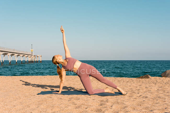 Mujer serena practicando yoga en Vasisthasana y equilibrándose en la rodilla en la orilla del mar en un día soleado - foto de stock