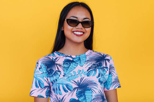 Happy Asian femelle dans des lunettes de soleil élégantes et t-shirt avec impression feuille tropicale en regardant la caméra sur fond jaune en studio — Photo de stock