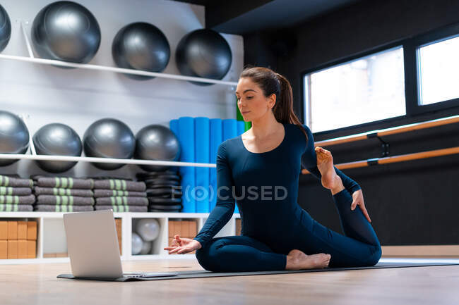 Full body of flexible young female yoga instructor doing One Legged King Pigeon variation pose with mudra gesture in front of laptop screen during online class in fitness studio — Stock Photo