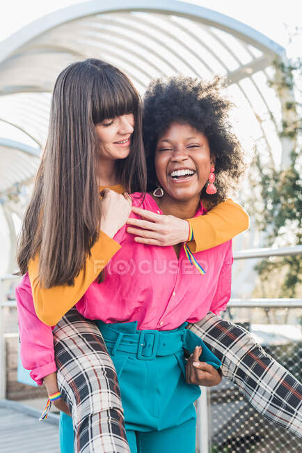 Sonriendo negro mujer piggybacking encantado lesbiana novia mientras se divierten en la ciudad en fin de semana y mirando a la cámara - foto de stock