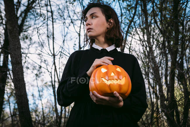 From below female in black dress standing with glowing pumpkin lantern in dark woods on Halloween looking away — Stock Photo
