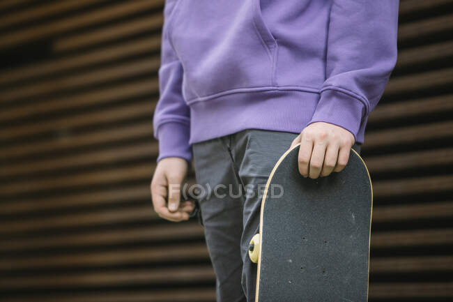 Unbekannter junger Mann in Freizeitkleidung mit Skateboard schaut in die Kamera, während er in der Nähe einer Mauer auf der Straße steht — Stockfoto