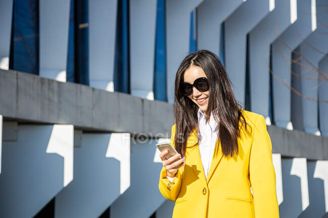 Азійська бізнес-жінка з жовтим пальто і розумний телефон ходити по вулиці з будівлею на задньому плані — стокове фото