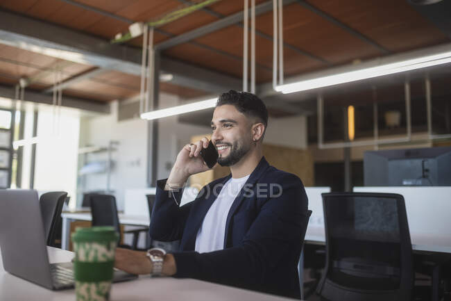 Веселый мужчина-предприниматель, говорящий по мобильному телефону и работающий на рабочем месте, сидя за столом с ноутбуком — стоковое фото