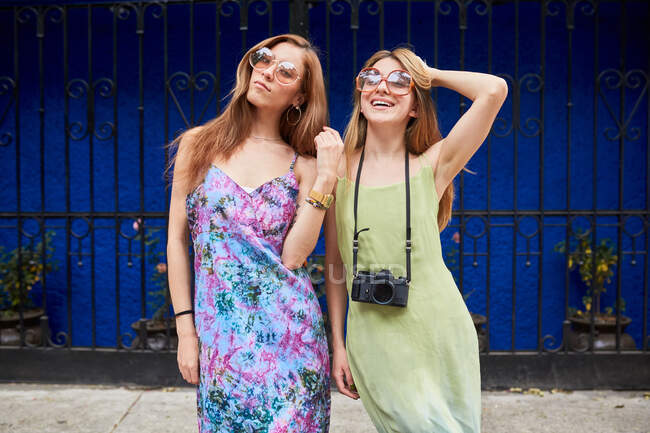 Young trendy female friends wearing summer dresses and sunglasses standing together in city street and looking at camera — Stock Photo