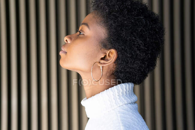 Side view of unemotional African American female in trendy sweater looking away against striped building wall on street — Stock Photo