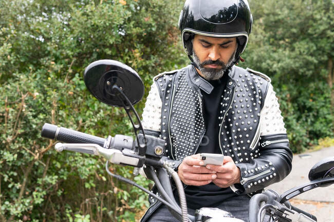Мужчина-байкер в модной кожаной куртке с заклепками и защитным шлемом просматривает телефон, сидя на мотоцикле, припаркованном возле пышного зеленого леса — стоковое фото