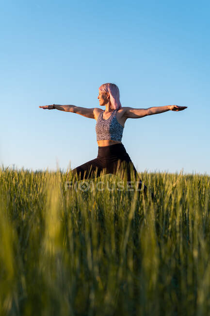 Adult female in sports clothes practicing yoga with outstretched arms on meadow under blue sky — Stock Photo