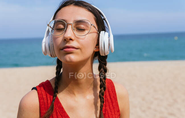 Транквіль жінка в навушниках слухає музику з закритими очима, насолоджуючись піснями на піщаному узбережжі в сонячний день влітку — стокове фото