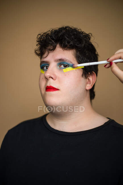 Estilista de cultivos con pincel aplicando maquillaje brillante en los párpados de los hombres queer mirando a la cámara - foto de stock
