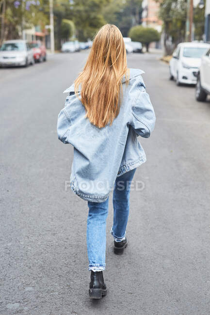 Задний вид молодой женщины в солнцезащитных очках и джинсовой одежде, смеющейся на улице, наслаждаясь выходными и глядя в камеру — стоковое фото