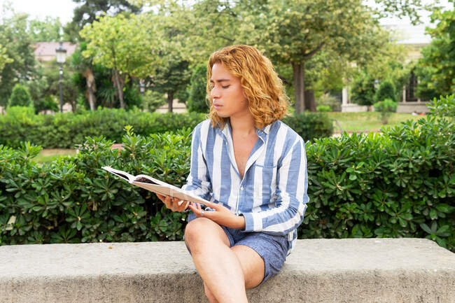Homme immobile aux cheveux longs assis sur un banc dans le jardin et lisant une histoire intéressante dans un livre pendant le week-end — Photo de stock