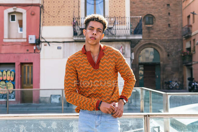 Молодой кудрявый парень в стильной разноцветной полосатой рубашке стоит на улице и задумчиво смотрит в камеру. — стоковое фото