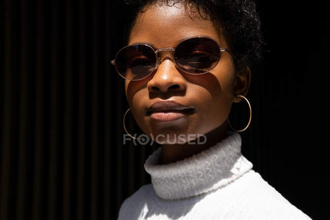 Молода афроамериканська жінка в стильному светрі і сонцезахисних окулярах дивиться на камеру, стоячи при яскравому світлі на чорному тлі. — стокове фото