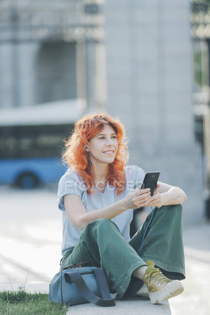 Вид збоку весела руда жінка сидить на вулиці і обмінюється повідомленнями в соціальних мережах на мобільний телефон під час прослуховування музики — стокове фото