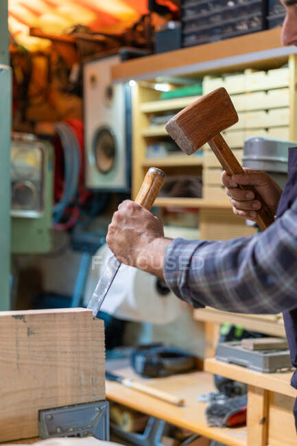 Side view of crop male master using hammer and chisel while creating wooden detail in carpentry workshop — Stock Photo