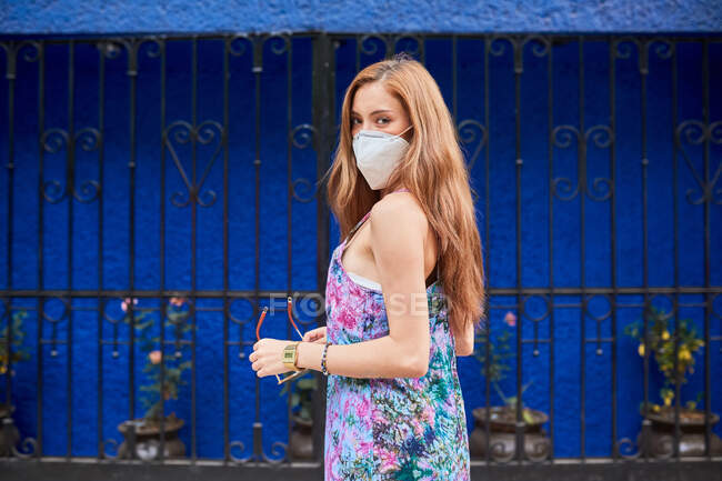 Вид збоку жінки в захисній масці і сукні, що стоїть на вулиці влітку під час епідемії коронавірусу — стокове фото