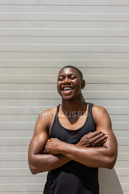 Ajuste hombre afroamericano de pie con los brazos cruzados cerca de la pared de metal en la calle y riendo con los ojos cerrados en el día soleado - foto de stock