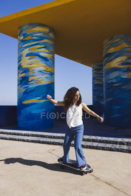 Full body of female in casual clothes riding skateboard on sunny street near colorful painted column during training — Stock Photo