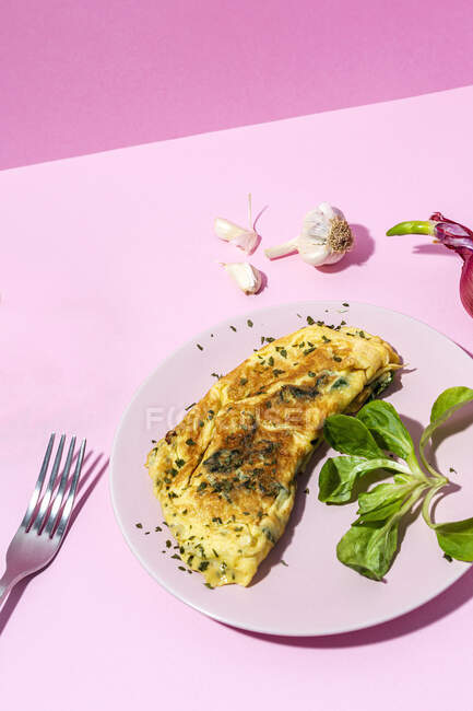 Tasty omelette on plate against fresh parsley sprigs and red onion with garlic cloves on pink background — Stock Photo