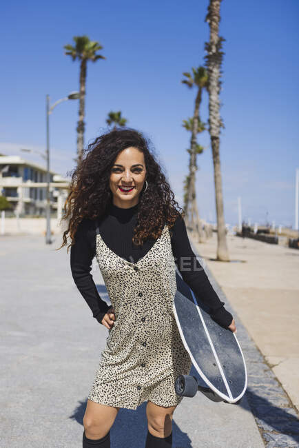 Femme joyeuse avec planche à roulettes dans les mains regardant la caméra tout en se tenant près de la côte de sable — Photo de stock