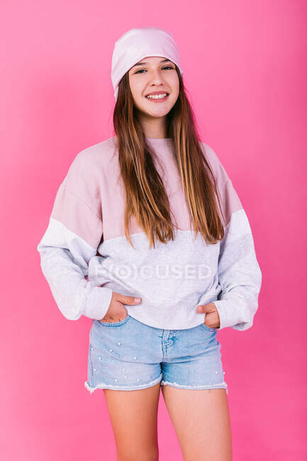 Contenu adolescent femme en foulard avec les cheveux bruns pour le concept de cancer en regardant la caméra sur fond rose avec les mains dans la poche — Photo de stock