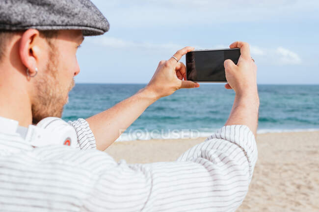 Vista lateral de un joven y elegante viajero barbudo tomando fotos de paisajes marinos en el teléfono móvil mientras pasa el día de verano en la playa de arena - foto de stock