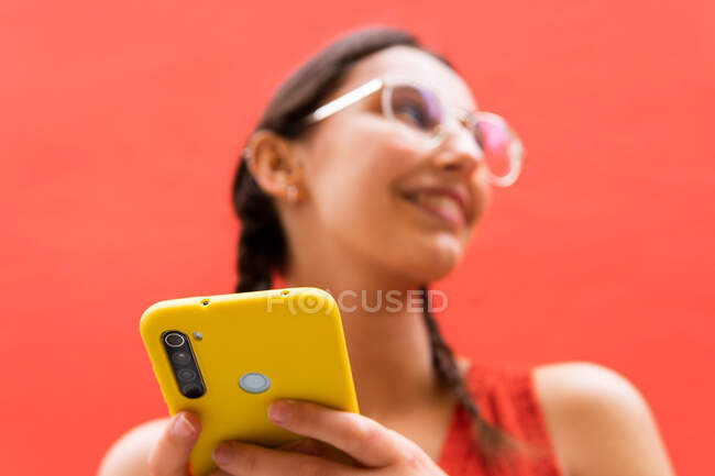 Снизу веселая молодая женщина в косичках прическа просматривает на смартфоне стоя глядя в сторону на красном фоне на улице — стоковое фото