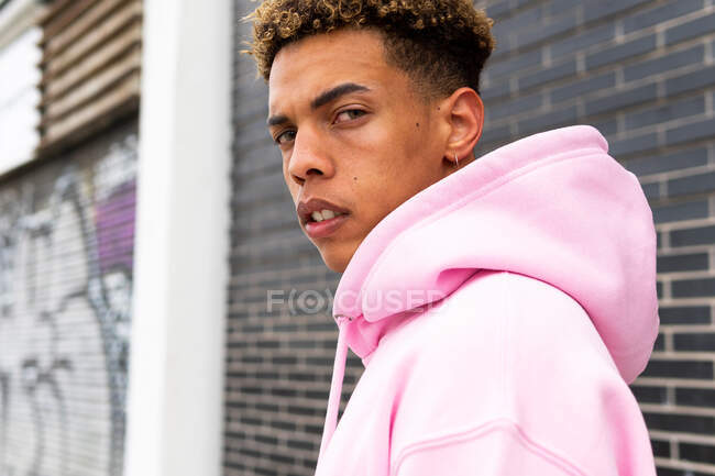 Side view of confident serious young curly haired hipster guy in pink hoodie looking at camera against brick wall — Stock Photo