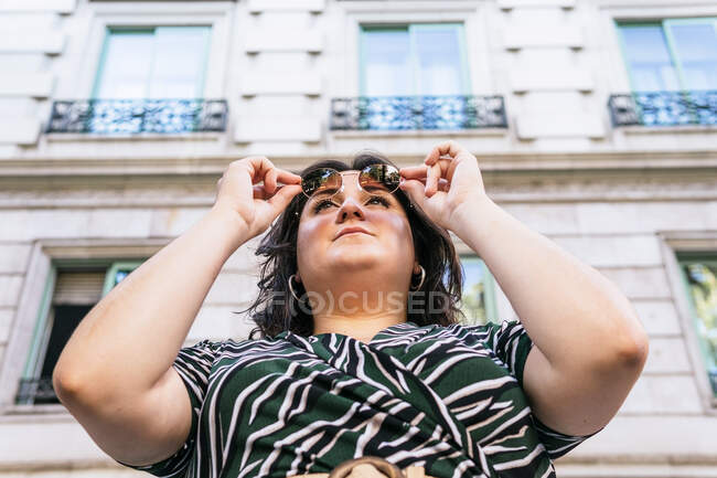 Низький кут впевненості молодої пишної жінки в стильній сукні з геометричним принтом і модними сонцезахисними окулярами, які дивляться далеко, стоячи біля кам'яної міської будівлі в літній день — стокове фото