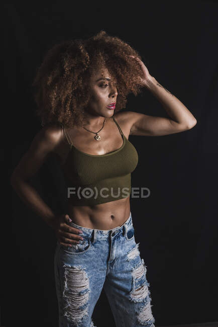 Cabelo encaracolado afro-americano fêmea em top de cultura na moda e jeans de pé com a mão na cintura em fundo preto no estúdio — Fotografia de Stock