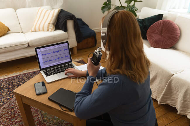 Vista posterior de una mujer irreconocible en el podcast de grabación de auriculares mientras habla en micrófono y usa el portátil en casa - foto de stock