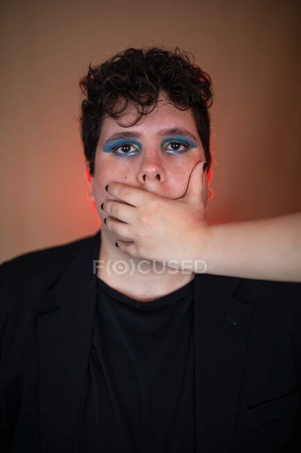 Homme sans émotion avec un maquillage lumineux avec la bouche couverte par la culture femelle regardant la caméra — Photo de stock
