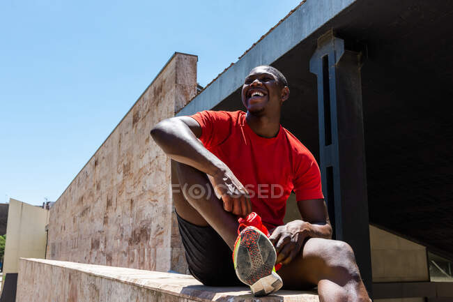 Low angle of smiling African American male athlete in activewear sitting with bottle of water on stone border in street during training and looking away — Stock Photo