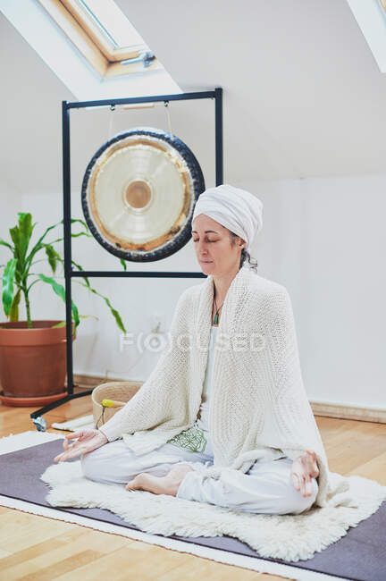 Mujer madura con los ojos cerrados sentada con las piernas cruzadas sobre una alfombra esponjosa mientras practica yoga en casa - foto de stock