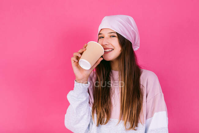 Frohe Teenagerin mit Kopftuch für Krebskonzept mit Pappbecher Heißgetränk zum Mitnehmen vor laufender Kamera — Stockfoto