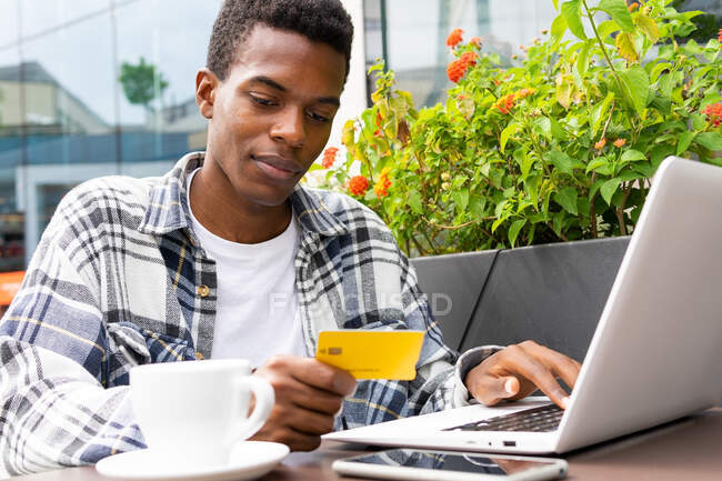 Konzentrierter afroamerikanischer Mann bezahlt für Bestellung mit Plastikkarte, während er Laptop beim Online-Einkauf im Straßencafé benutzt — Stockfoto