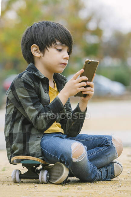 Vista lateral del adorable chico con ropa casual navegando por el teléfono celular mientras está sentado en el monopatín en el cruce peatonal en la ciudad - foto de stock