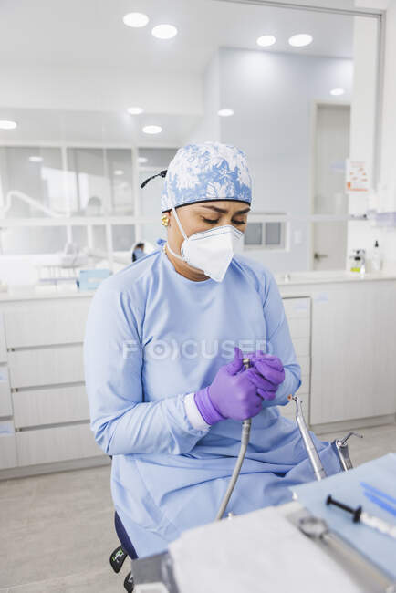 Dentista feminino étnico grave em máscara e uniforme médico com luvas trabalhando na máquina de perfuração na clínica — Fotografia de Stock
