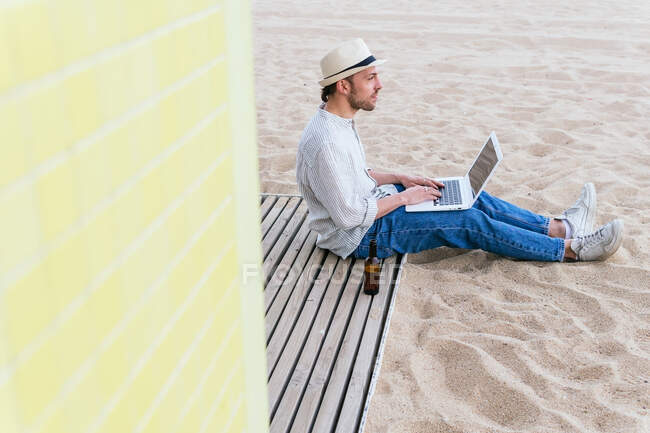 Full body side view of young male blogger in stylish wear and hat sitting with bottle of beer on sandy beach and typing on laptop during summer holidays on seashore — Stock Photo