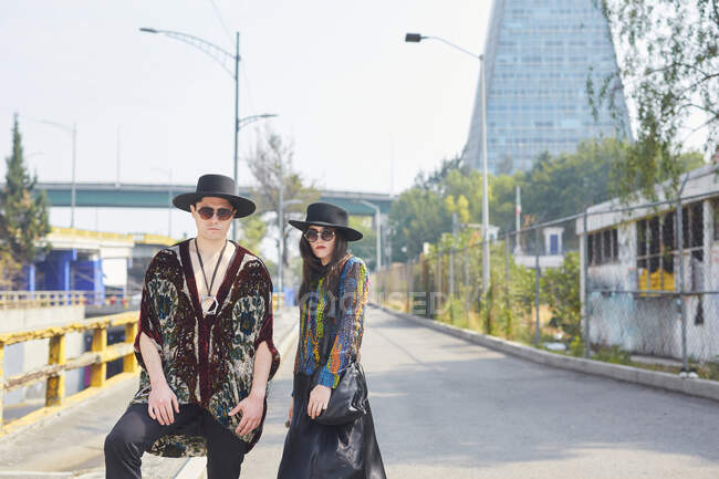 Couple élégant sérieux en vêtements hipster debout regardant la caméra le long de la rue en été — Photo de stock