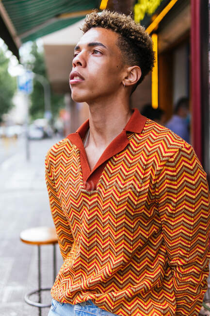 Молодой этнический кудрявый парень в стильной разноцветной полосатой рубашке стоит на улице и задумчиво смотрит в сторону. — стоковое фото