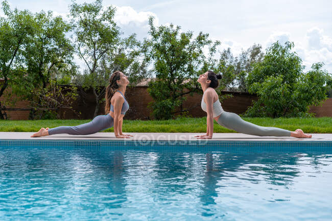 Side view of women with eyes closed in sportswear performing Toe Squat pose while practicing yoga in Cobra pose between swimming pool and lawn — Stock Photo